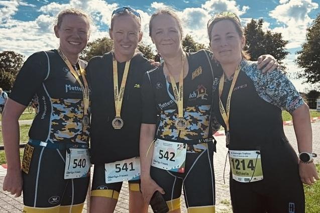 Vier Frauen im Sportoutfit mit Medaillen um den Hals posieren l&auml;chelnd nach einem Wettkampf auf einem Veranstaltungsgel&auml;nde.