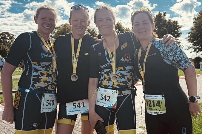 Vier Frauen im Sportoutfit mit Medaillen um den Hals posieren lächelnd nach einem Wettkampf auf einem Veranstaltungsgelände.