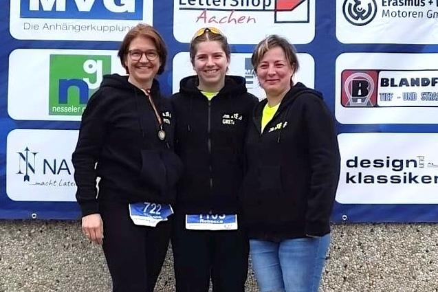 Drei Frauen in Sportkleidung posieren l&auml;chelnd vor einer Sponsorenwand nach einem Wettkampf.