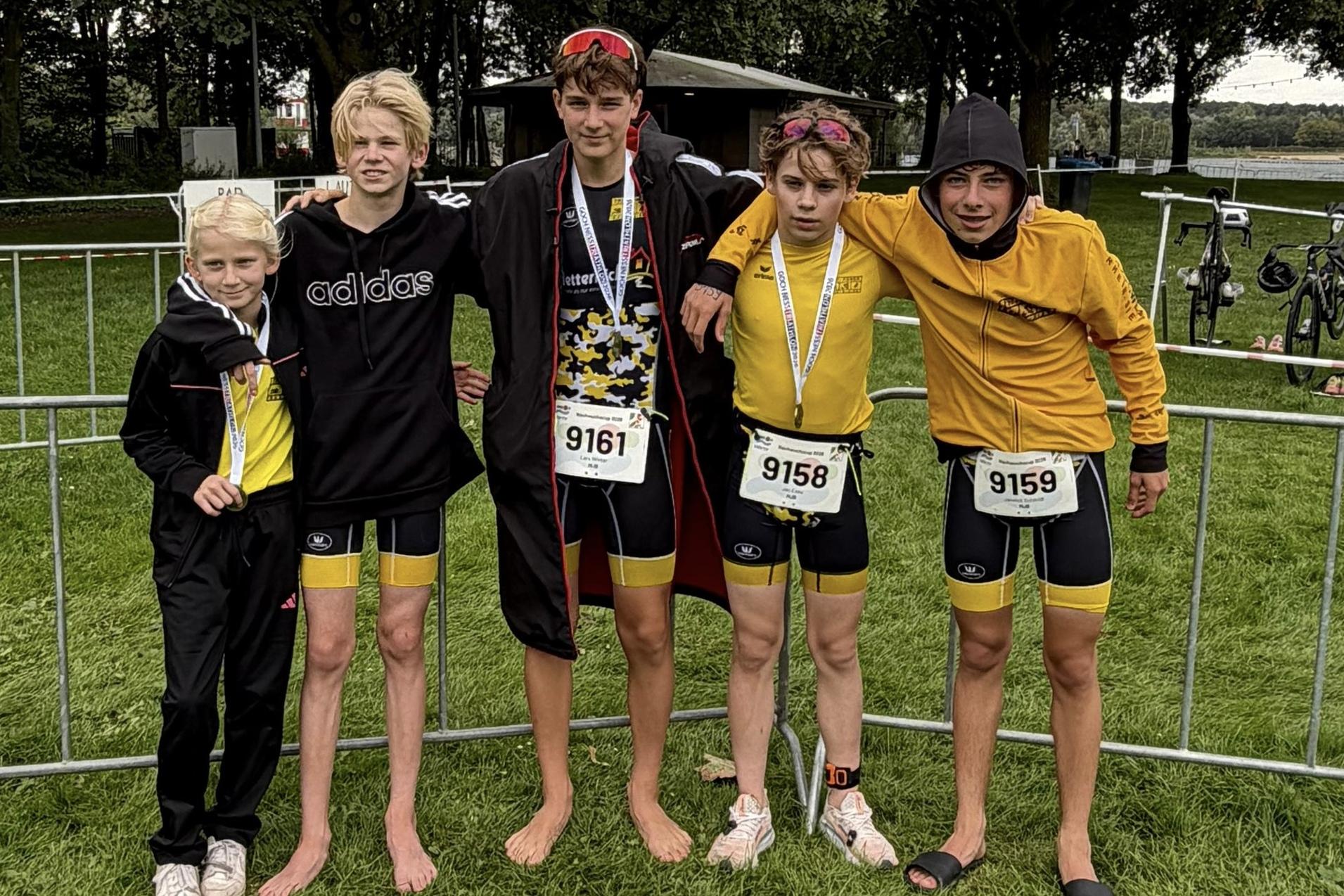 F&uuml;nf junge Triathleten in Sportkleidung stehen l&auml;chelnd an einem gesperrten Bereich nach einem Wettkampf.