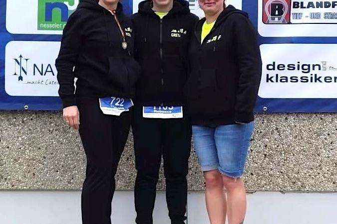 Drei Frauen in Sportkleidung posieren l&auml;chelnd vor einer Sponsorenwand nach einem Wettkampf.