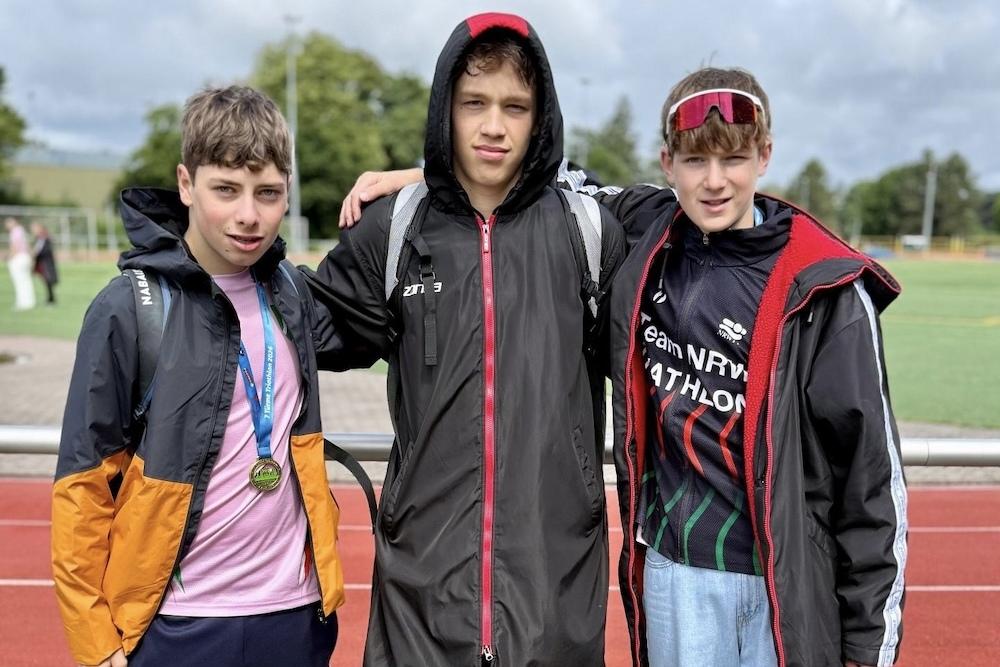 Drei junge Sportler stehen arm in arm auf einem Sportfeld, tragen l&auml;ngere Jacken und Blick in die Kamera.