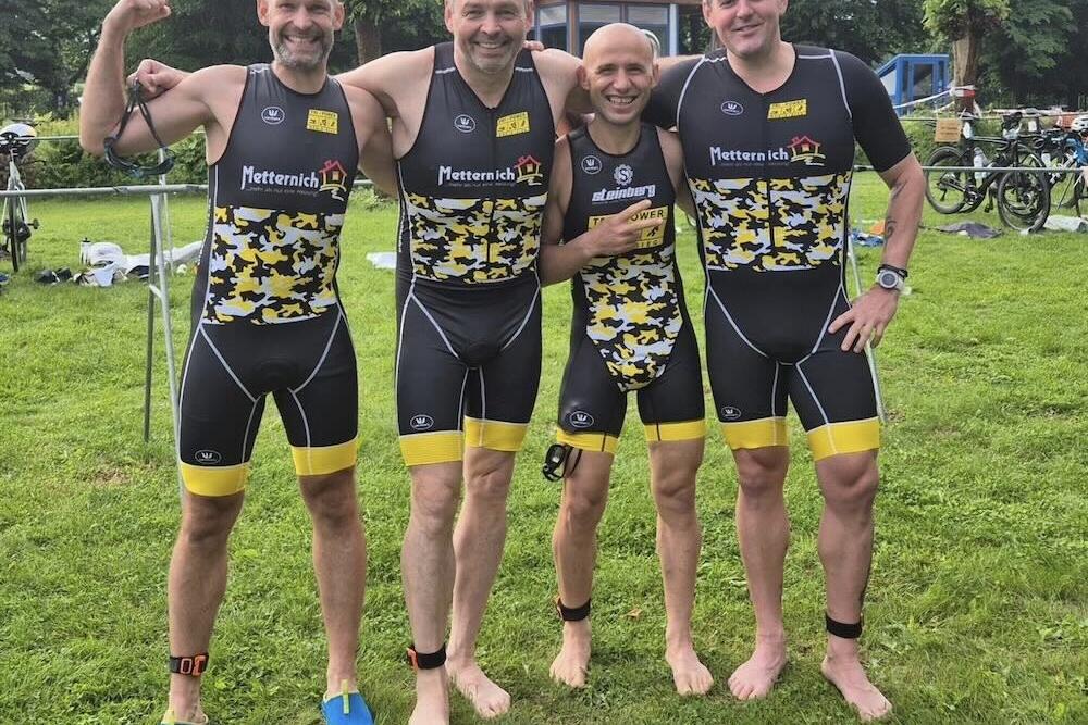 Vier M&auml;nner in sportlichen Triathlon-Anz&uuml;gen posieren l&auml;chelnd auf einer gr&uuml;nen Wiese, umgeben von Fahrr&auml;dern.
