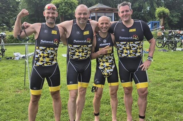 Vier M&auml;nner in sportlichen Triathlon-Anz&uuml;gen posieren l&auml;chelnd auf einer gr&uuml;nen Wiese, umgeben von Fahrr&auml;dern.