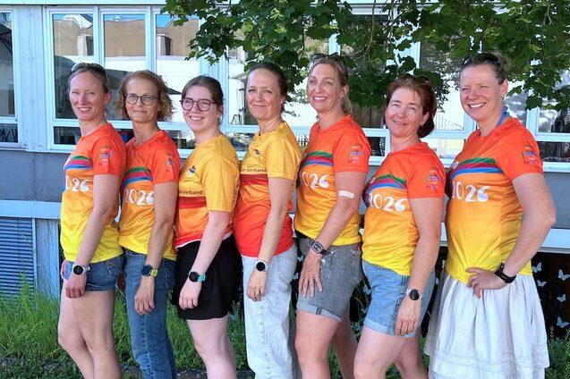 Gruppe von Frauen in bunten Sportshirts vor einem Geb&auml;ude, l&auml;chelnd und nebeneinander aufgestellt.