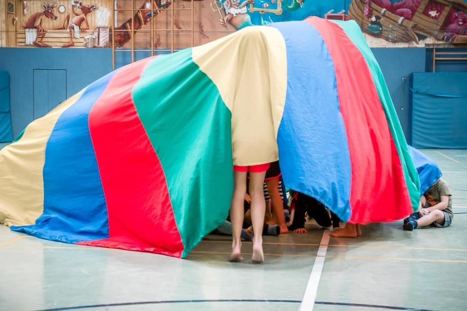 Kinder spielen unter einem bunter, farbigen Fallschirm in einer Turnhalle.