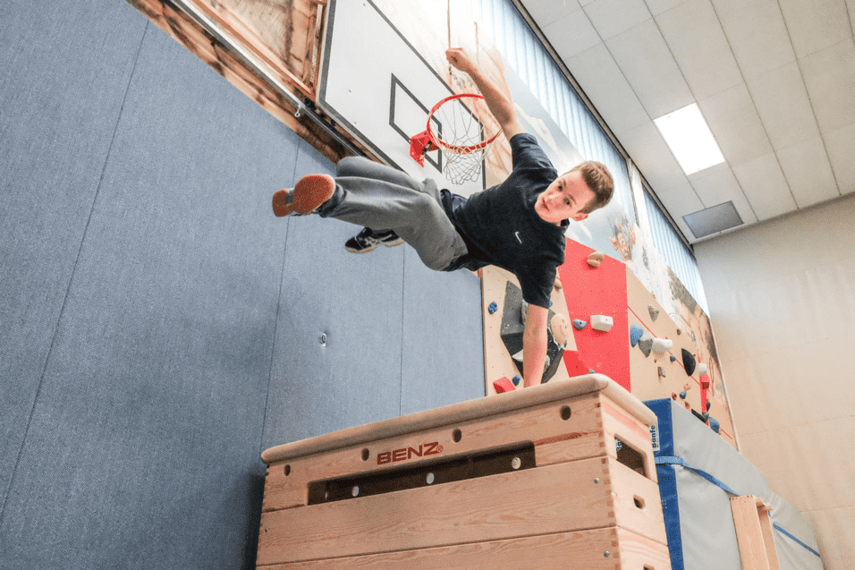 Jugendlicher springt akrobatisch &uuml;ber eine Holzbank in einer Sporthalle mit Kletterwand im Hintergrund.