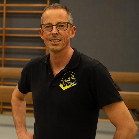 Trainer steht l&auml;chelnd vor einem Schwebebalken, w&auml;hrend ein Kind auf dem Balken balanciert.