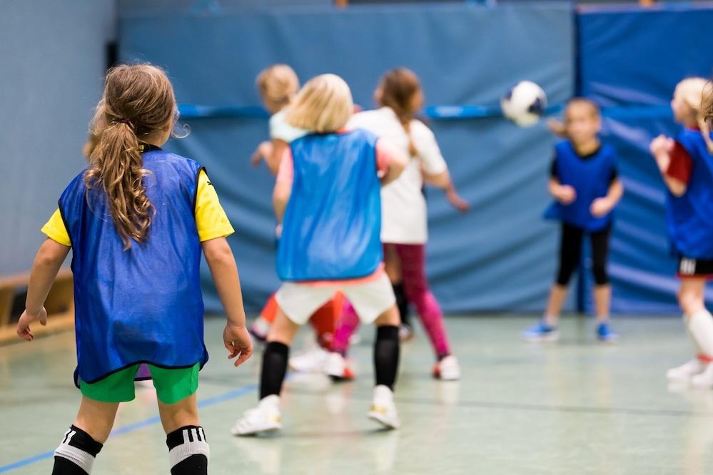 M&auml;dchen in Trainingsanz&uuml;gen spielen Fu&szlig;ball in einer Sporthalle, w&auml;hrend der Ball in die Gruppe fliegt.