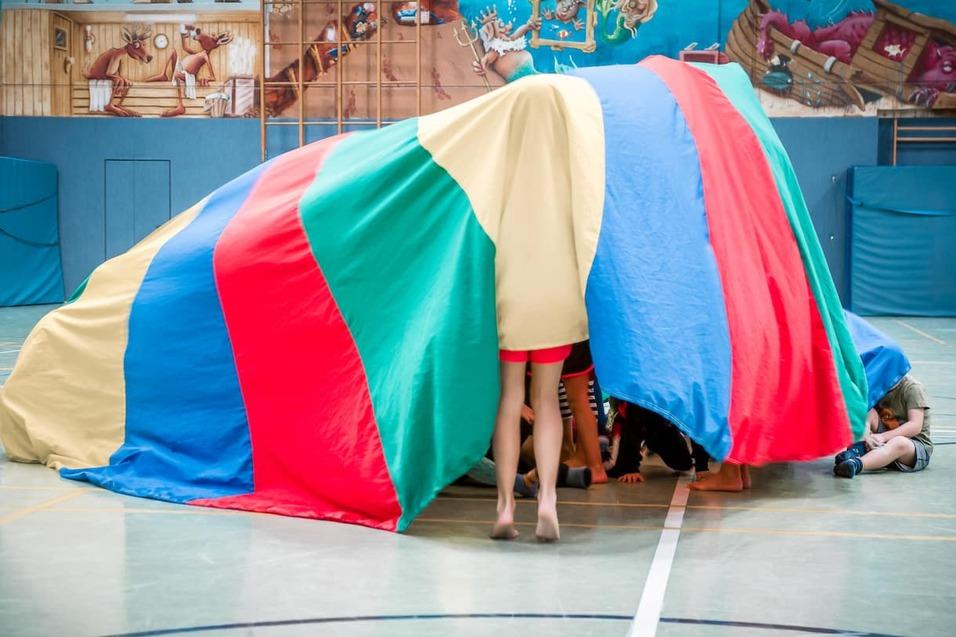 Kinder spielen unter einem bunter, farbigen Fallschirm in einer Turnhalle.