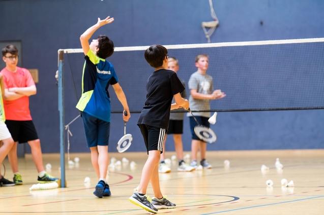 Zwei Kinder spielen Badminton in einer Sporthalle, w&auml;hrend weitere Kinder im Hintergrund beobachten. Federn liegen auf dem Boden.