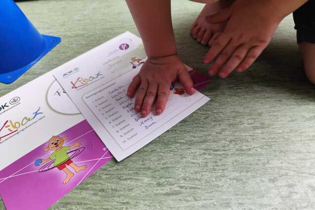 Kind legt seine Hand auf ein Arbeitsblatt mit einem bunten Design auf dem Boden. Im Hintergrund ist ein blauer Kegel sichtbar.