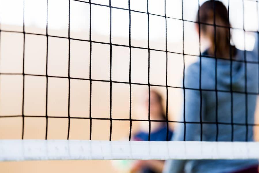 Nahaufnahme eines Volleyballnetzes mit verschwommenen Spielern im Hintergrund in einer Sporthalle.