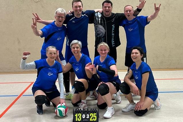Gruppenselfie von acht Volleyballspielern in blauen Trikots, jubelnd vor einer Wand mit Punkteanzeige (13:25).