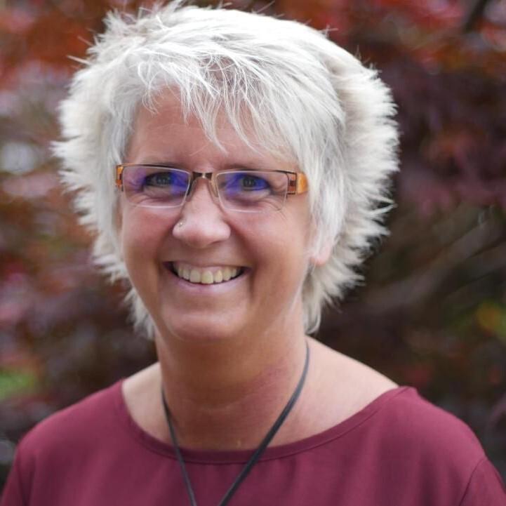 Porträt einer lächelnden Frau mit kurzen, grauen Haaren, Brille und einem burgunderfarbenen Top, vor einer unscharfen, natürlichen Kulisse.