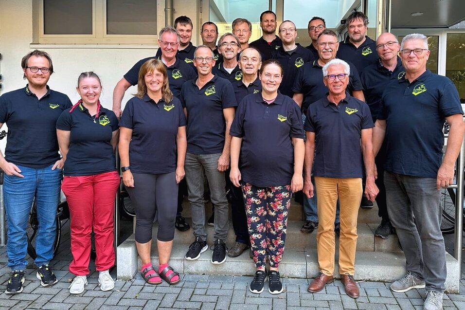 Gruppe von 20 Personen in schwarzen Shirts mit Logo, stehend vor einem Gebäude, während sie lächeln.