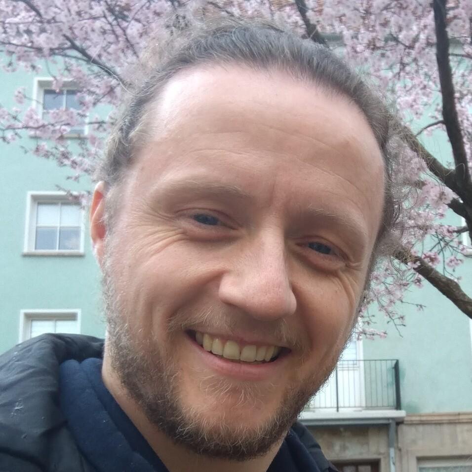 Lächelnder Mann mit kurzer Haarfrisur und Bart, vor blühenden Kirschbäumen und einer grünen Wand.
