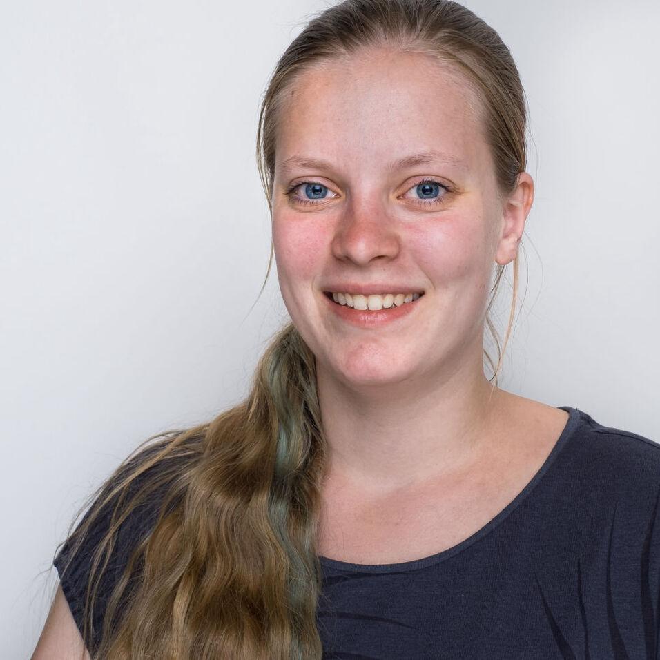 Lächelnde Frau mit langen, blonden Haaren und blauen Augen vor einem hellen Hintergrund.