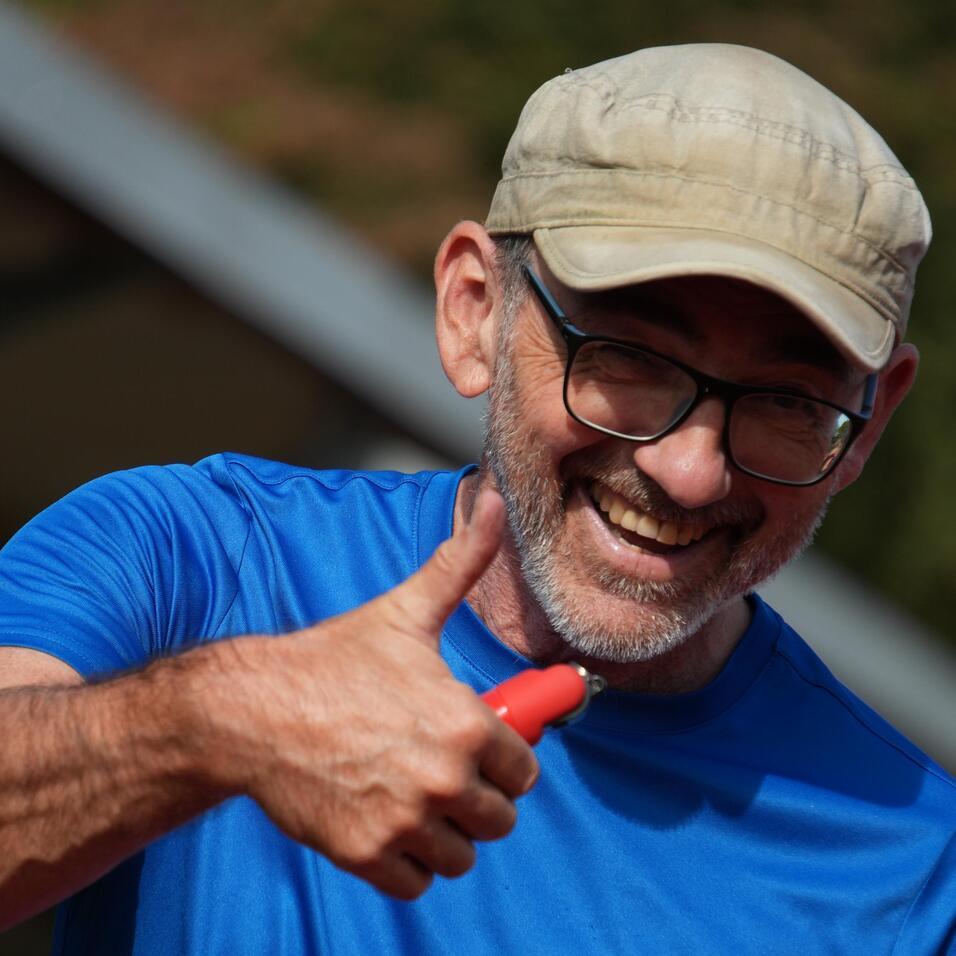 Lächelnder Mann mit Brille und Kappe zeigt Daumen hoch, trägt ein blaues T-Shirt und hat ein Namensschild mit „Richard“.