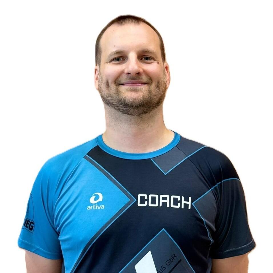 L&auml;chelnder Mann in einem blauen Sportshirt mit Aufschrift &bdquo;COACH&ldquo; vor einem einfarbigen Hintergrund.