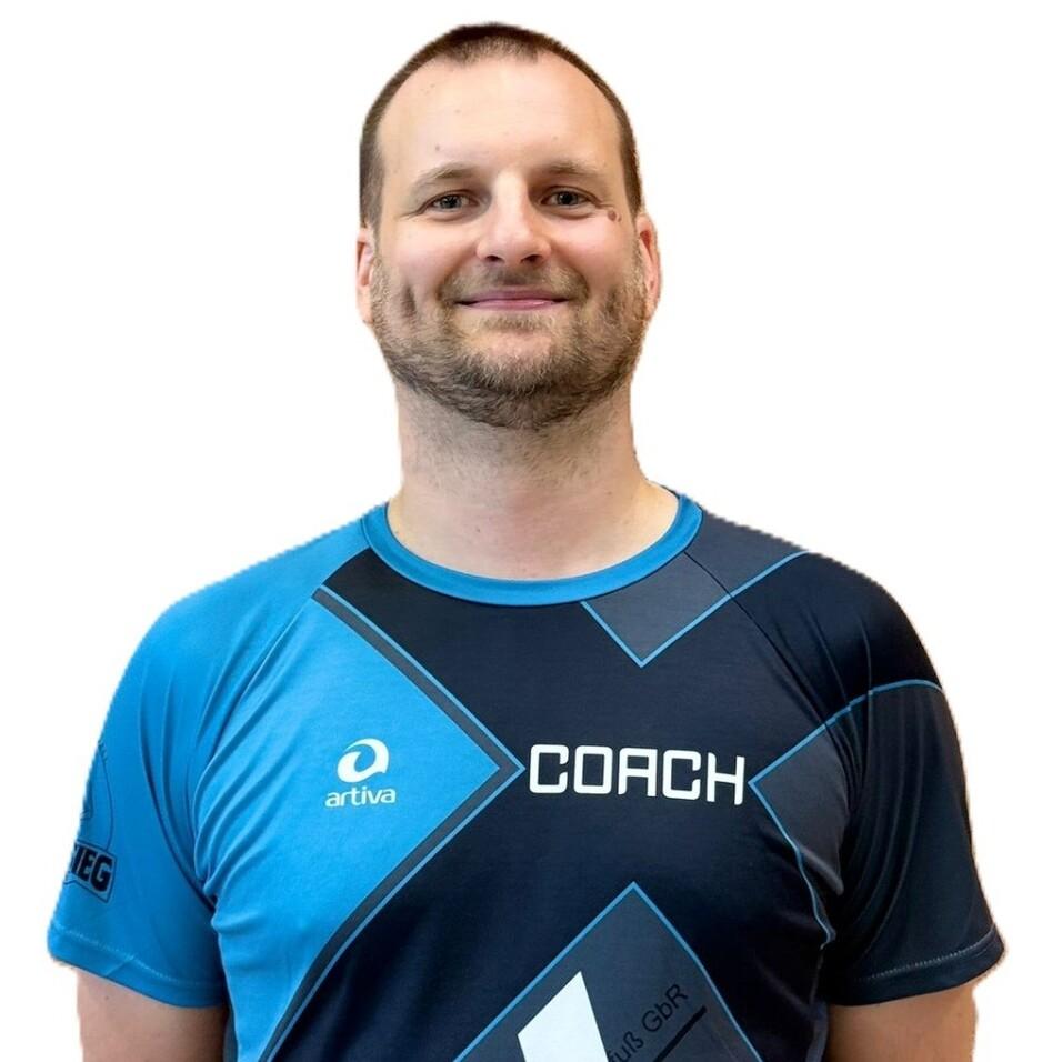 Lächelnder Mann in einem blauen Sportshirt mit Aufschrift „COACH“ vor einem einfarbigen Hintergrund.