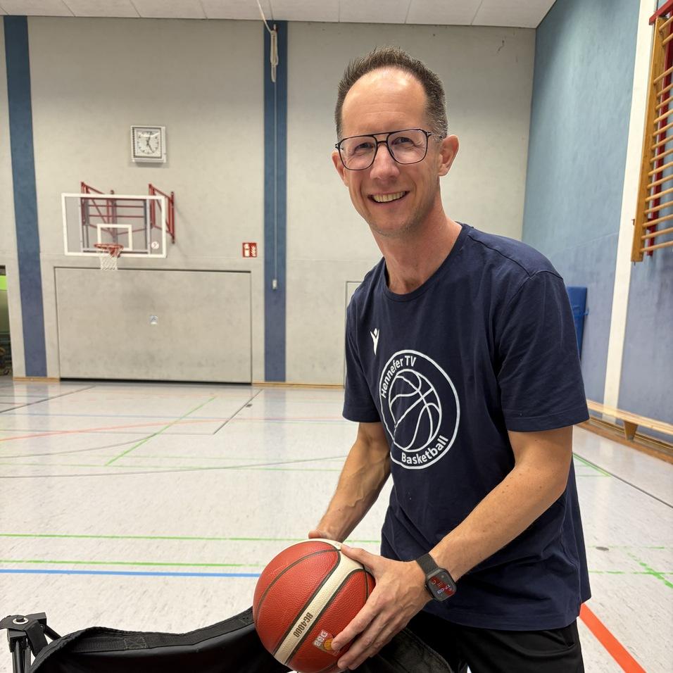 Mann mit Brille in Basketball-T-Shirt hält einen Basketball über einem Netz mit mehreren Bällen in einer Sporthalle.