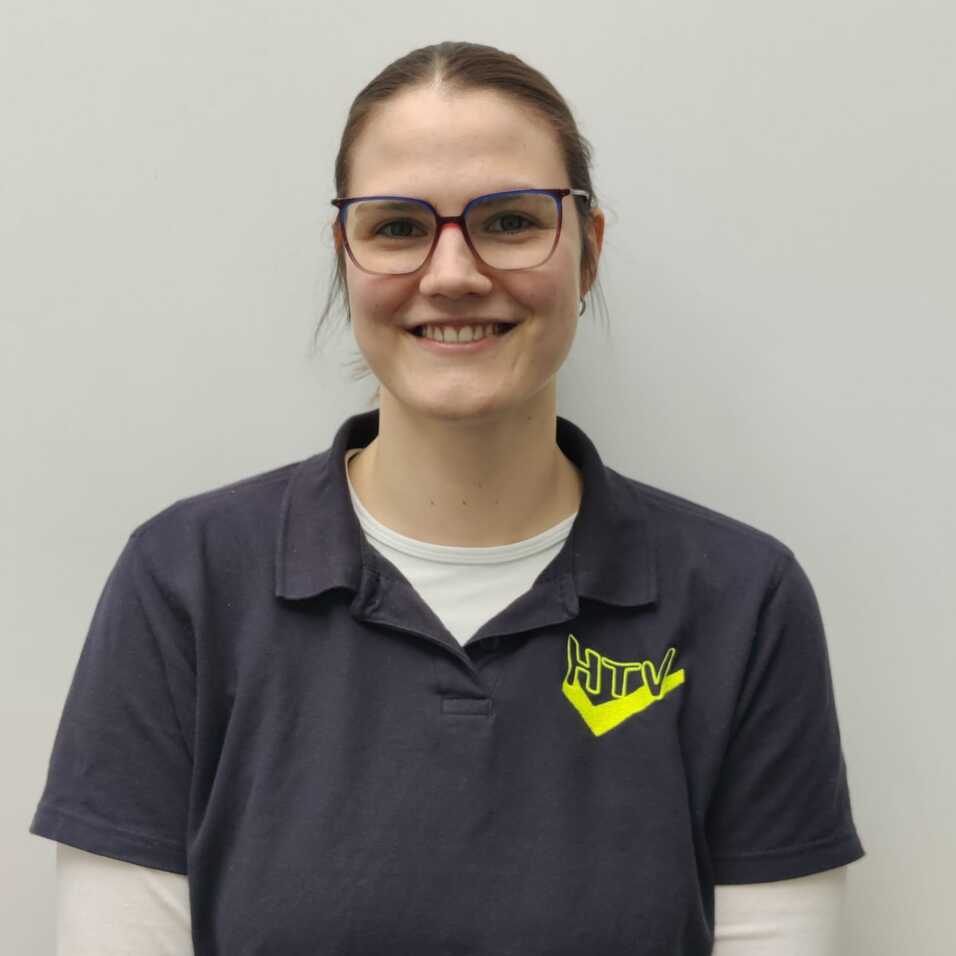 Porträt einer lächelnden Frau mit Brille, die ein graues Polo-Shirt mit gelbem Logo trägt, vor einer hellen Wand.