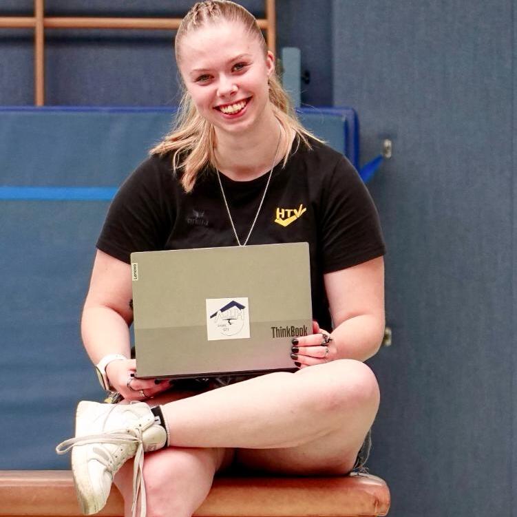 Junge Frau sitzt auf einem Turnbock und l&auml;chelt, w&auml;hrend sie ein Laptop mit Aufkleber h&auml;lt. Turnhalle im Hintergrund.