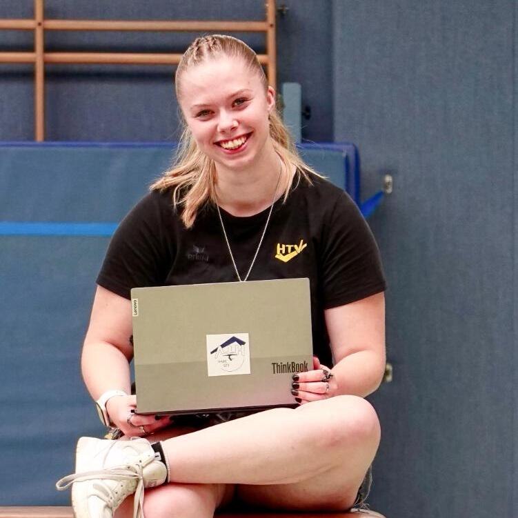 Junge Frau sitzt auf einem Turnbock und l&auml;chelt, w&auml;hrend sie ein Laptop mit Aufkleber h&auml;lt. Turnhalle im Hintergrund.