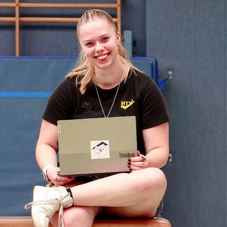 Junge Frau sitzt auf einem Turnbock und l&auml;chelt, w&auml;hrend sie ein Laptop mit Aufkleber h&auml;lt. Turnhalle im Hintergrund.