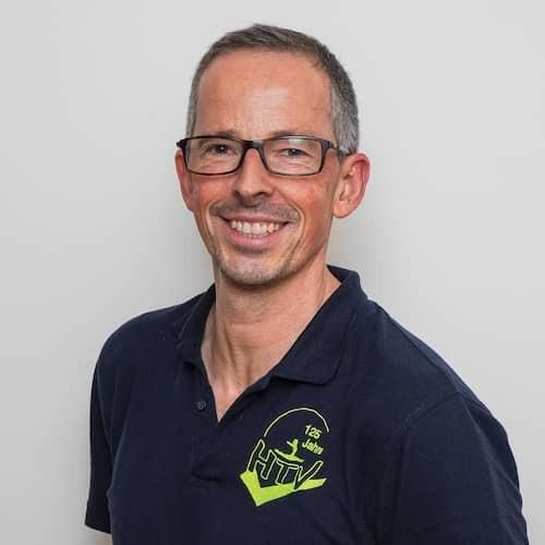 Porträt eines lächelnden Mannes mit Brille, der ein dunkelblaues Shirt mit grünem Logo trägt. Hintergrund grau.