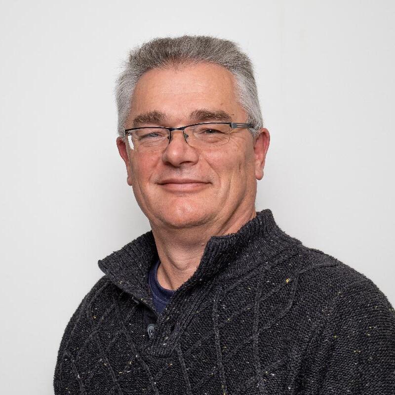 Porträt eines Mannes mit Brille in einem grauen Pullover vor einem hellen Hintergrund. Er lächelt leicht in die Kamera.