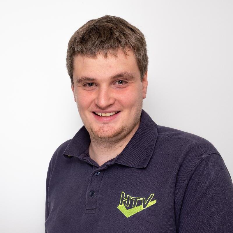 Portrait eines lächelnden Mannes in einem dunkelblauen Polohemd mit Logo vor neutralem Hintergrund.