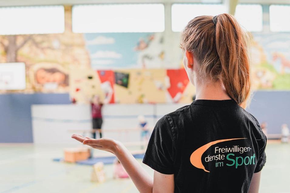 Eine Person mit langen Haaren in einem schwarzen T-Shirt mit gelbem Logo steht in einer Sporthalle und beobachtet Kinder.
