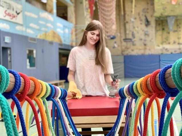 Junge Frau steht an einer Bank, umgeben von bunten Seilen in einer Sporthalle mit Kletterwand im Hintergrund.