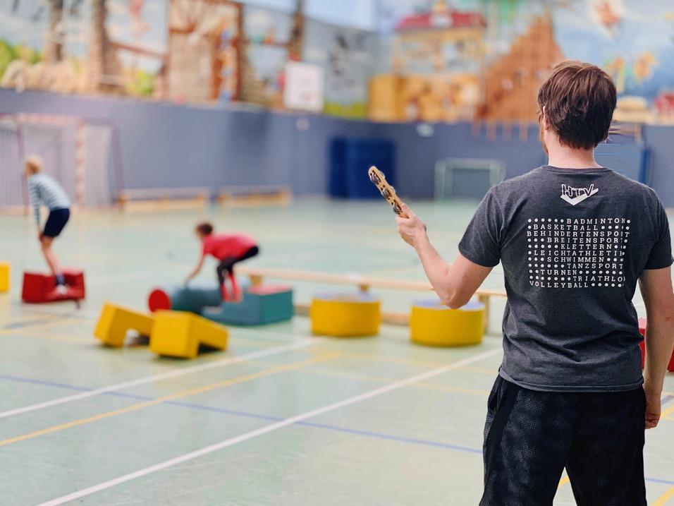 Ein Mann mit einem Schläger beobachtet Kinder, die auf verschiedenen Spielgeräten in einer Sporthalle balancieren.