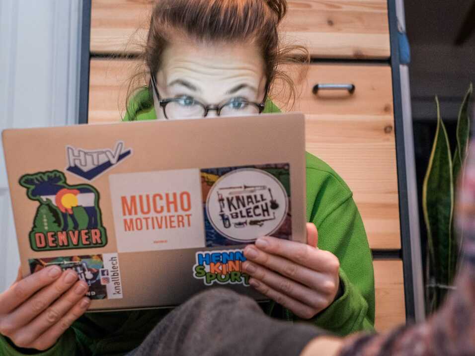 Eine Person mit Brille sitzt auf dem Boden und schaut auf einen Laptop, der mit verschiedenen Stickern beklebt ist.