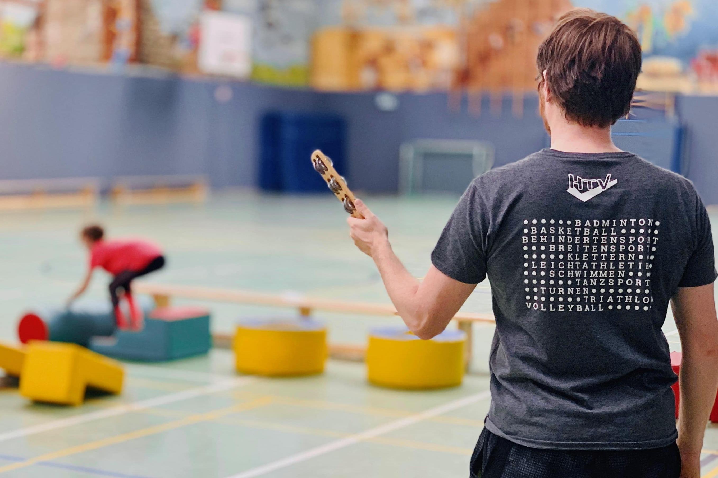 Ein Mann mit einem Schl&auml;ger beobachtet Kinder, die auf verschiedenen Spielger&auml;ten in einer Sporthalle balancieren.