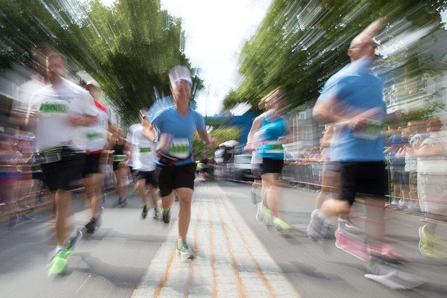 Schwenkaufnahme von laufenden Teilnehmern eines Straßenlaufes, umgeben von Zuschauern und grünen Bäumen.