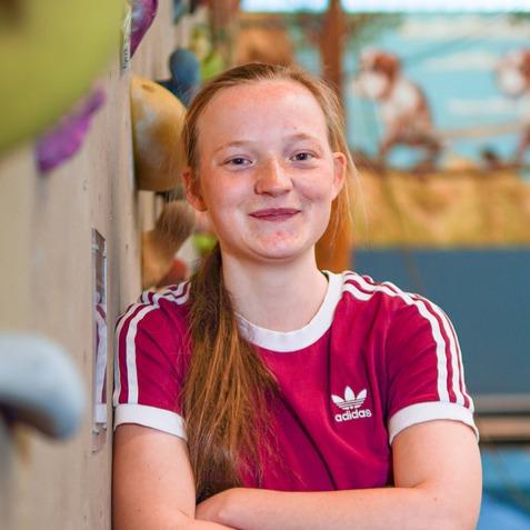 Mädchen mit langem Haar in rotem Sportshirt lehnt an einer Kletterwand in einer Kletterhalle.