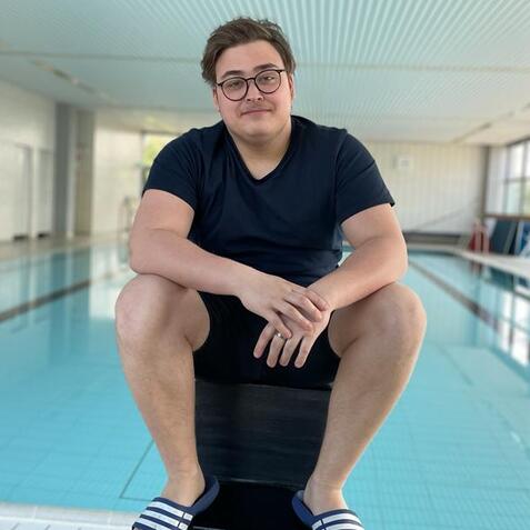 Junger Mann sitzt am Rand eines Schwimmbeckens, trägt ein schwarzes T-Shirt und Schlappen, Blick auf das Wasser gerichtet.