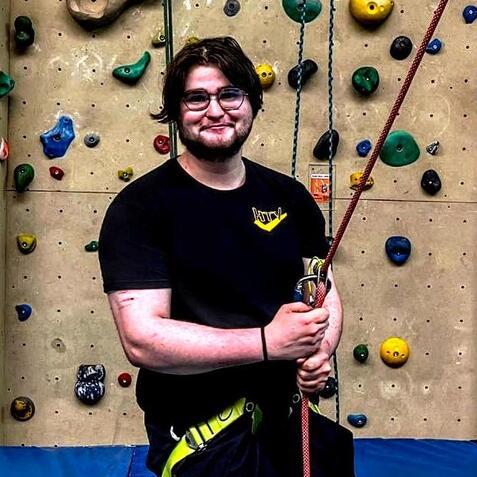 Junger Mann steht mit Sicherungsseil in der Hand vor einer Kletterwand mit bunten Griffen in einer Kletterhalle.