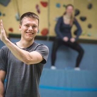 Lächelnder junger Mann in Sportkleidung mit erhobener Hand, während eine Frau hinter ihm Seilspringen übt.
