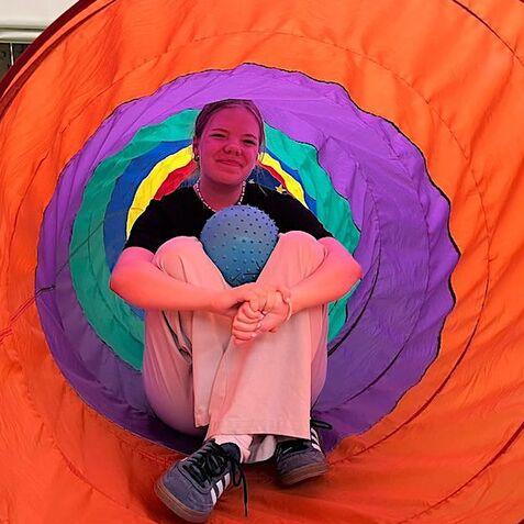Mädchen sitzt in einem bunten Tunnelspielzeug und hält einen blauen Ball in einer Turnhalle.