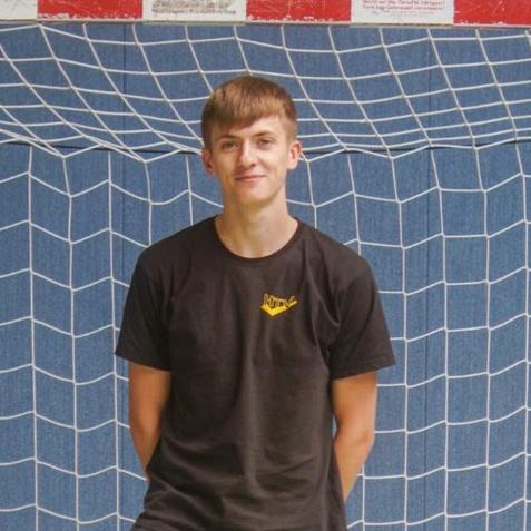 Junger Mann steht in einem Hallenfußballtor, hinter ihm ein Netz und blauer Wandhintergrund. Er trägt ein schwarzes T-Shirt.