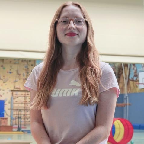 Junge Frau mit langen roten Haaren sitzt auf einer Schaukel in einer Sporthalle mit Kletterwand im Hintergrund.