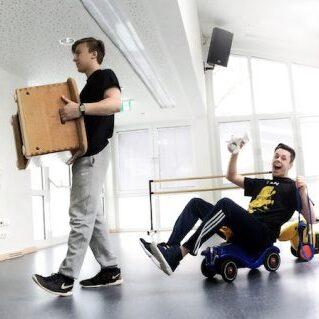 Fünf Personen in einem Raum, jeweils in unterschiedlichen Positionen, eine trägt einen Karton, eine sitzt auf einem Spielzeugfahrzeug.