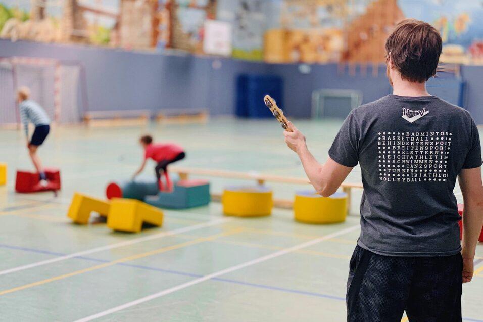 Junge mit Badmintonschl&auml;ger beobachtet Kinder, die auf bunten Kletterelementen in einer Sporthalle spielen.