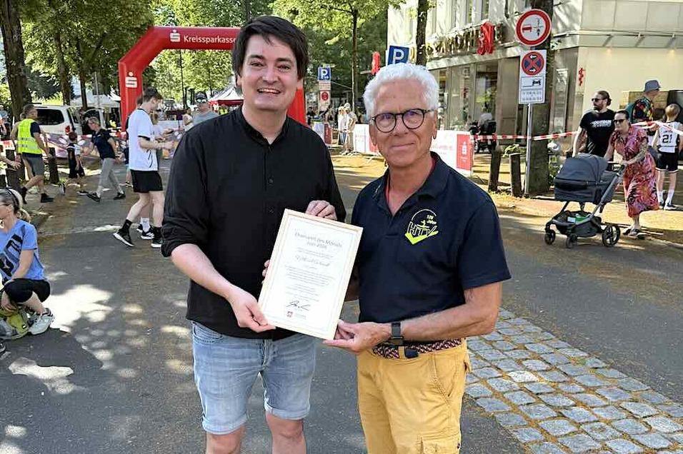 Zwei Männer stehen auf einer Straße, einer hält ein Urkunde, der andere lächelt neben ihm. Im Hintergrund eine belebte Kulisse.