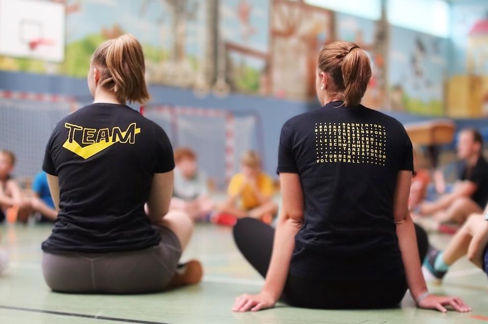 Zwei Personen mit T-Shirts, eins mit „TEAM“ und das andere mit verschiedenen Sportarten, sitzen im Kreis auf dem Boden.